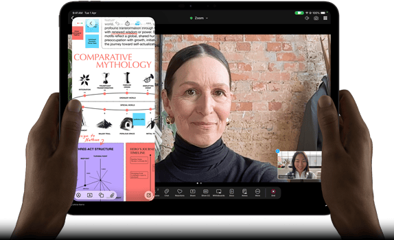 iPad Pro, front exterior, landscape orientation, space black color, rounded corners, black display bezel, hands holding left and right sides, display showing a FaceTime conversation being live translated, and an educational app showing a presentation of "Comparative Mythology" with text, diagrams, and handwritten notes